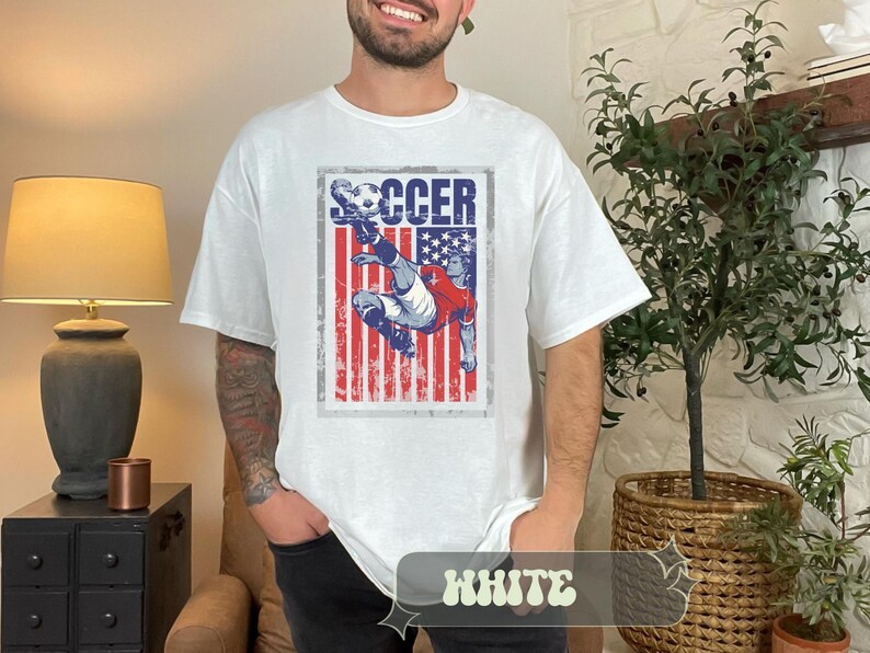 May include: White t-shirt with a distressed graphic featuring a soccer player kicking a ball, set against a backdrop of the American flag. The word "SOCCER" is printed above the image. The shirt is a casual, unisex style.