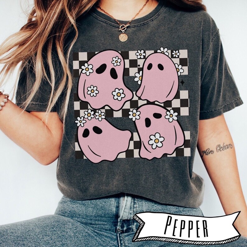 May include: Dark gray t-shirt featuring a graphic of four pink ghosts with daisy crowns on a black and white checkered background. The ghosts have black eyes and are smiling. The word "PEPPER" is printed at the bottom.