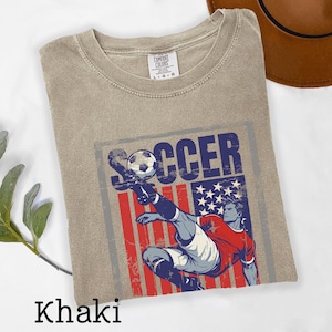 May include: Khaki-colored t-shirt featuring a graphic of a soccer player kicking a ball, with an American flag design. The word "SOCCER" is printed above the player. The shirt has a crew neck and short sleeves.