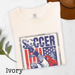 May include: Ivory-colored t-shirt with a graphic featuring a soccer player kicking a ball. The design incorporates the American flag with red and blue elements, and the word "SOCCER". The shirt is a Comfort Colors brand.