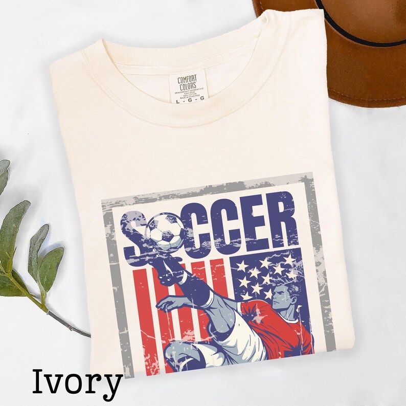 May include: Ivory-colored t-shirt with a graphic featuring a soccer player kicking a ball. The design incorporates the American flag with red and blue elements, and the word "SOCCER". The shirt is a Comfort Colors brand.