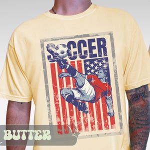 May include: Butter-colored t-shirt featuring a distressed graphic of a soccer player kicking a ball. The graphic incorporates the American flag with the word "SOCCER" in bold letters. The shirt has short sleeves.