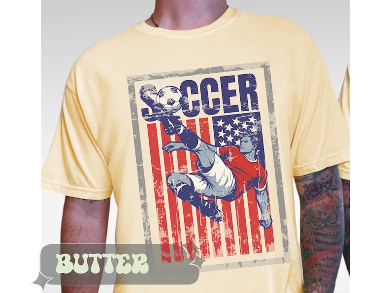 May include: Butter-colored t-shirt featuring a distressed graphic of a soccer player kicking a ball. The graphic incorporates the American flag with the word "SOCCER" in bold letters. The shirt has short sleeves.