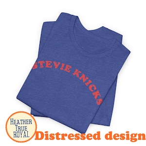 Puede incluir: Una camiseta de color azul real jaspeado con el texto STEVIE NICKS impreso en una fuente roja desgastada en el pecho. La prenda cuenta con cuello redondo y mangas cortas, mostrando una textura de tela con un acabado suave y jaspeado.