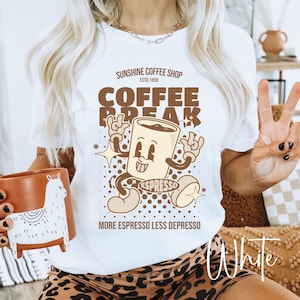 May include: White t-shirt with a brown cartoon coffee cup graphic. The graphic says "Sunshine Coffee Shop Est. 1958 Coffee Break" and "Espresso" on the cup. The text below the graphic says "More Espresso Less Depresso".