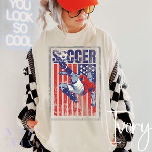 May include: Ivory-colored t-shirt featuring a distressed graphic of a soccer player kicking a ball, set against a red, white, and blue American flag design. The word "SOCCER" is printed above the graphic. The shirt is worn by a person wearing an orange cap.