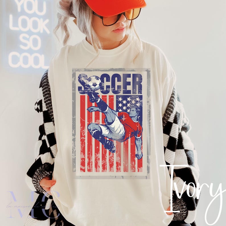 May include: Ivory-colored t-shirt featuring a distressed graphic of a soccer player kicking a ball, set against a red, white, and blue American flag design. The word "SOCCER" is printed above the graphic. The shirt is worn by a person wearing an orange cap.