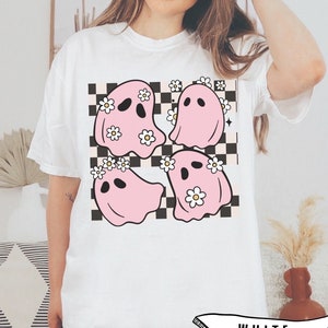 May include: White t-shirt featuring a graphic of four pink ghosts with daisy crowns on a black and white checkered background. The word "WHITE" is printed on a banner at the bottom of the shirt.