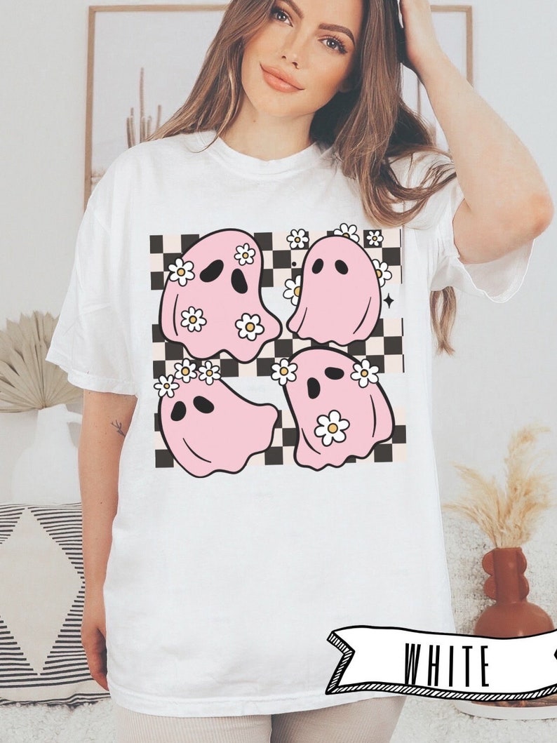 May include: White t-shirt featuring a graphic of four pink ghosts with daisy crowns on a black and white checkered background. The word "WHITE" is printed on a banner at the bottom of the shirt.