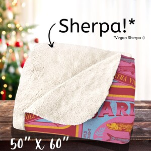 May include: A pink and blue throw blanket with a vintage-style fish can design. The blanket is 50 inches by 60 inches and has a soft, plush sherpa lining.