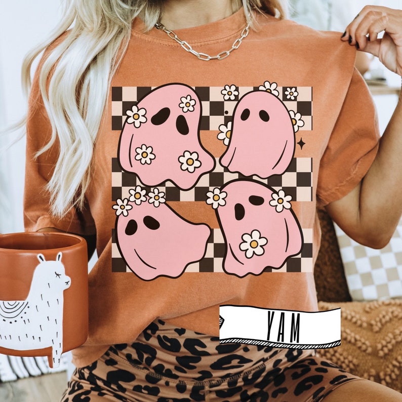 May include: Orange-colored t-shirt featuring a graphic of four pink ghosts with daisy crowns on a checkered background. The ghosts have black eyes and smiles. The shirt is paired with leopard print shorts.