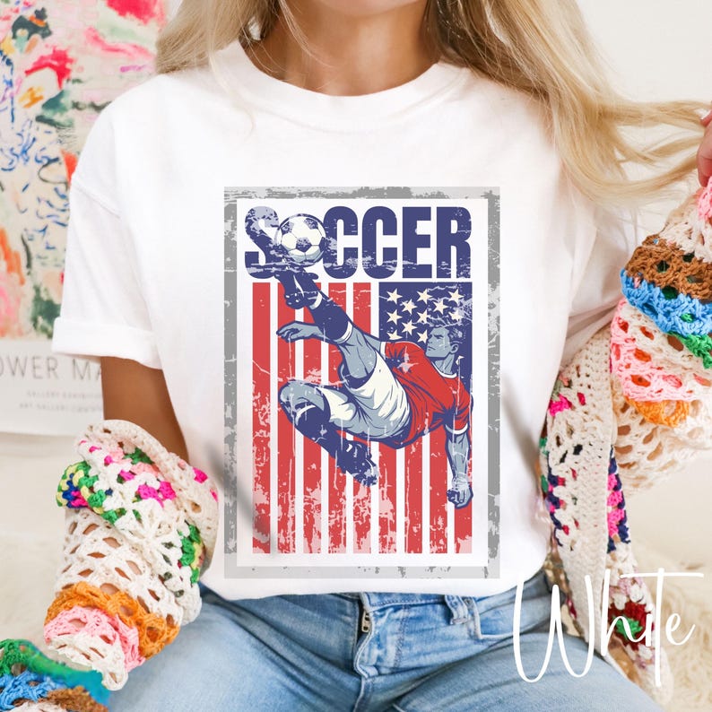 May include: White t-shirt featuring a distressed graphic of a soccer player kicking a ball. The design incorporates the American flag with red and white stripes and a blue field of stars. The word "SOCCER" is printed above the player.