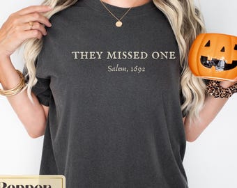 May include: A dark gray t-shirt with the text "They Missed One Salem, 1692" printed on the front. The shirt is being modeled by a person holding a jack-o-lantern.