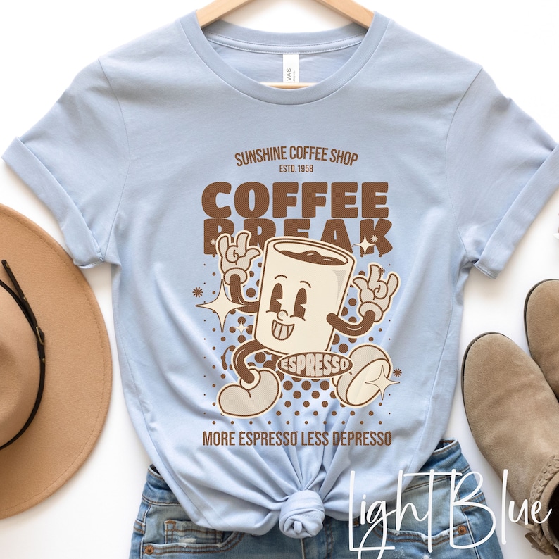 May include: Light blue t-shirt with a cartoon coffee cup graphic. The coffee cup is wearing a brown hat and has a smiling face. The text on the shirt reads "Sunshine Coffee Shop Est. 1958 Coffee Break More Espresso Less Depresso".