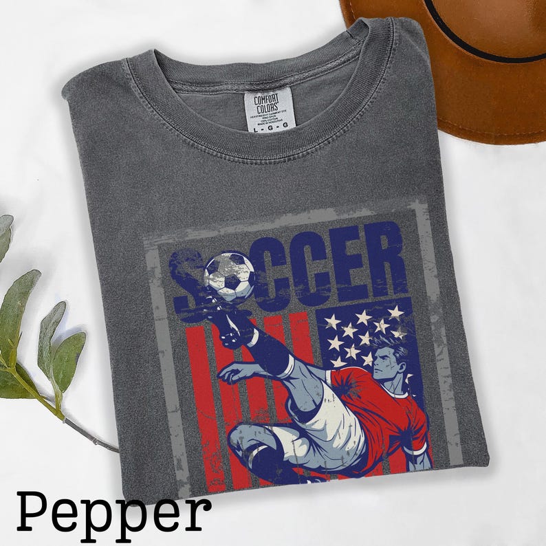 May include: Pepper-colored t-shirt with a soccer-themed graphic. The design features a soccer player kicking a ball, with a red, white, and blue flag design and the word "SOCCER". The shirt is a Comfort Colors brand.