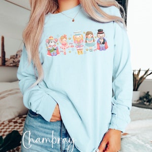 May include: A light blue long-sleeved shirt with a graphic of a Nutcracker and other characters. The graphic includes a mouse, a ballerina, a nutcracker, a princess, and a magician. The graphic is in a watercolor style and features a light blue background with gold stars.