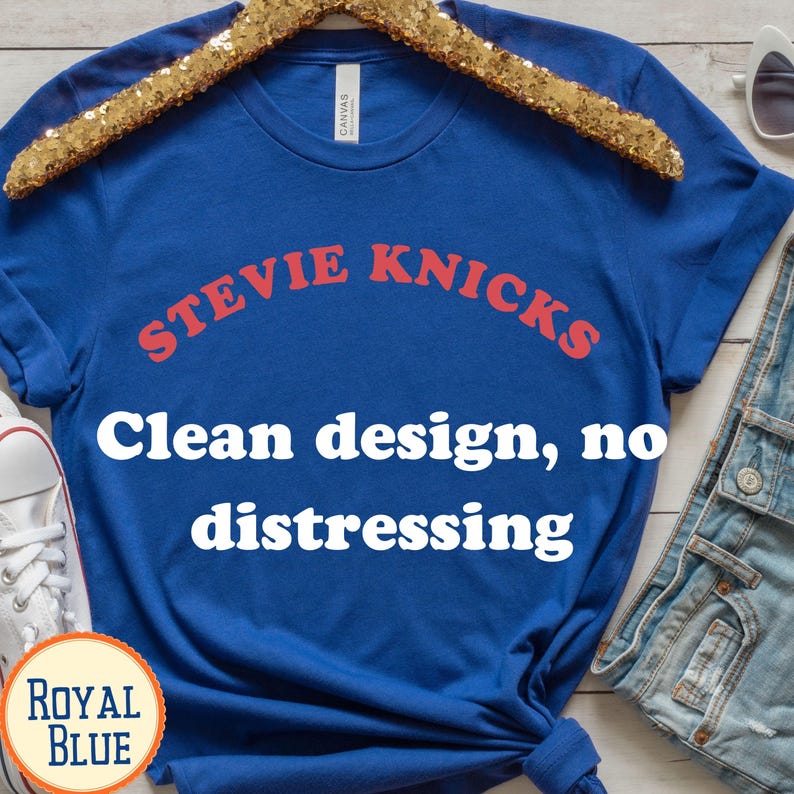 Puede incluir: Una camiseta de manga corta color azul real con el texto rojo STEVIE KNICKS arqueado en el pecho. Debajo del nombre, el texto blanco dice Clean design, no distressing. La camiseta se muestra en una percha de lentejuelas doradas con etiqueta.