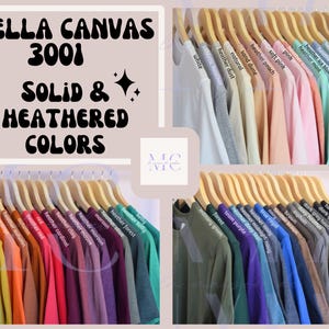 Puede incluir: Una colecci&oacute;n de camisetas Bella Canvas 3001 en colores s&oacute;lidos y jaspeados en perchas de madera. La imagen incluye etiquetas de texto para cada color, incluyendo blanco, heather dust, sand dune, heather peach, rosa, heather sunset, heather mint, azul hielo, azul polvoriento, salvia, amarillo, mostaza, naranja, oto&ntilde;o, rojo, cardenal, arcilla, malva, granate, oliva, bosque, kelly, verde militar, morado, azul medianoche, azul real, marr&oacute;n, asfalto, gris oscuro y negro.