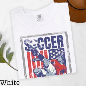 May include: White t-shirt with a distressed graphic featuring a soccer player kicking a ball, set against a backdrop of the American flag. The word "SOCCER" is printed above the player. The shirt is labeled "Comfort Colors".