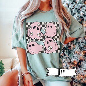 May include: Sage green t-shirt featuring a graphic of four pink ghosts with daisy crowns on a black and white checkered background. The word "BAY" is printed on a white banner at the bottom of the shirt.