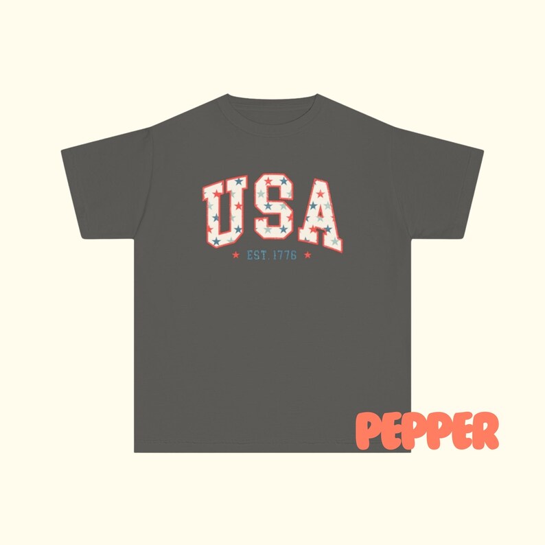May include: Dark gray t-shirt with the word "USA" in a vintage, collegiate-style font. The letters are filled with a pattern of red, white, and blue stars. Below the word "USA" is the text "EST. 1776" in a smaller font, with stars on either side.