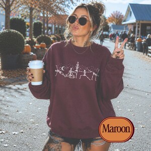 May include: A maroon long-sleeve crewneck sweatshirt featuring a white graphic print of five skeletons in various yoga poses, interspersed with small stars and a crescent moon. The sweatshirt has a ribbed collar, cuffs, and hemline.