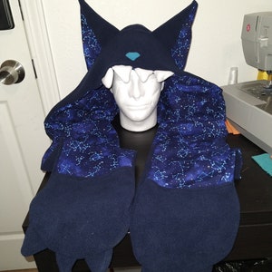 May include: A dark blue fleece hooded scarf featuring large pointed ears, a small teal nose, and white teeth along the forehead. The interior lining displays a vibrant blue and purple galaxy print with white star constellations throughout the fabric.