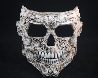 May include: A white skull mask with intricate carvings and a toothy grin. The mask is made of a smooth, textured material and has a slightly distressed look.