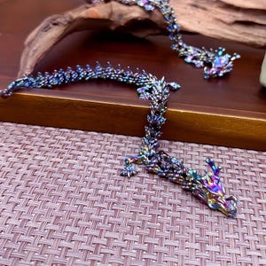 May include: A multi-colored iridescent dragon-shaped necklace made of metal. The necklace is on a woven surface.