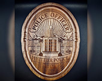 May include: An oval wooden plaque featuring a laser-engraved design of Whittier City Hall. The text reads POLICE OFFICER at the top, WHITTIER POLICE at the bottom, and 217 in a central shield. The wood grain is visible throughout the brown surface.
