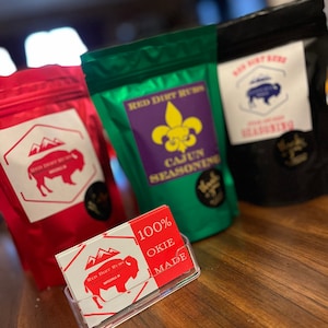 Peut inclure: Trois sachets d'assaisonnement au frottement de terre rouge, l'un avec une étiquette d'assaisonnement cajun, l'autre avec une étiquette d'assaisonnement BBQ et le dernier avec une étiquette simple. Une carte de visite avec le texte "100% OKIE MADE" est au premier plan.