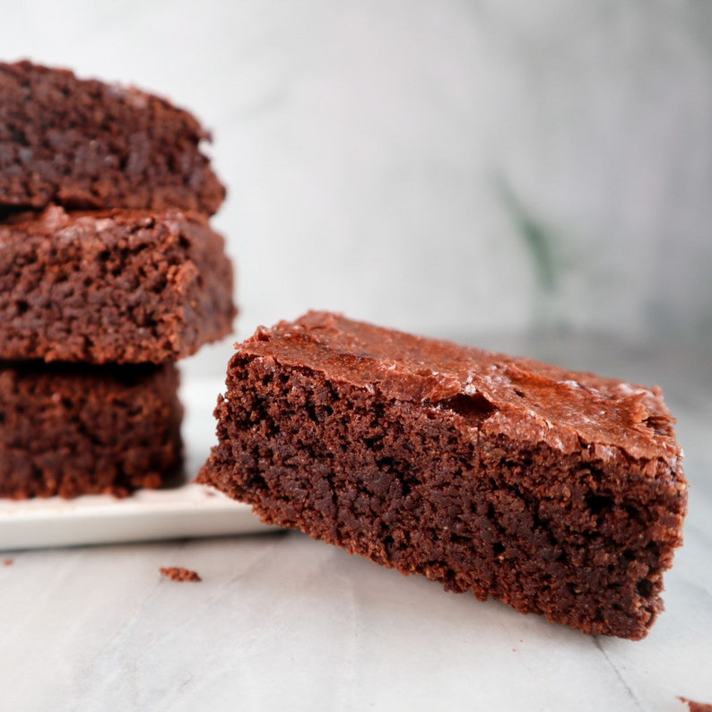 Puede incluir: Una pila de cuatro brownies de chocolate, con un brownie levantado de la pila e inclinado hacia un lado. Los brownies son de un color marr&oacute;n oscuro intenso y tienen una superficie ligeramente brillante.