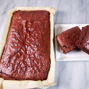 Puede incluir: Una bandeja grande de brownies de chocolate de un rico color marr&oacute;n oscuro. Los brownies est&aacute;n ligeramente agrietados en la superficie y tienen un acabado brillante. Dos brownies m&aacute;s peque&ntilde;os se muestran en un plato blanco.