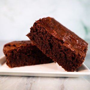 Puede incluir: Dos brownies de chocolate en un plato blanco. Los brownies son ricos y densos, con una parte superior ligeramente crujiente.