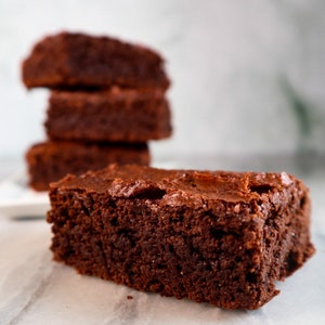 Puede incluir: Un primer plano de un brownie fudgy, con una pila de tres brownies m&aacute;s en el fondo. Los brownies son de un color marr&oacute;n oscuro intenso y tienen una superficie ligeramente brillante.