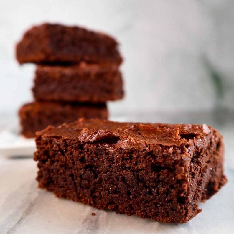 Puede incluir: Un primer plano de un brownie fudgy, con una pila de tres brownies m&aacute;s en el fondo. Los brownies son de un color marr&oacute;n oscuro intenso y tienen una superficie ligeramente brillante.