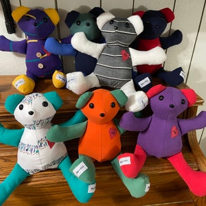 K&ouml;nnte beinhalten: Eine Sammlung von acht Pl&uuml;schb&auml;ren in verschiedenen Farben und Mustern. Die B&auml;ren sind in einer Reihe auf einer Holzoberfl&auml;che angeordnet. Die B&auml;ren sind alle handgefertigt und haben einzigartige Designs.