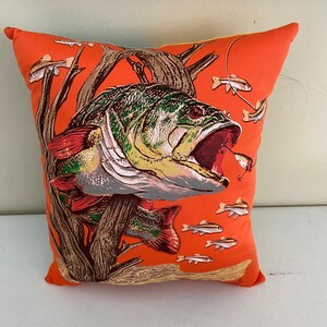 K&ouml;nnte beinhalten: Ein orangefarbener Dekokissen mit einer gro&szlig;en Illustration eines Barsches mit offenem Maul. Der Fisch ist gr&uuml;n, braun und orange mit einem roten Maul. Kleinere orangefarbene Fische schwimmen um den Barsch herum.