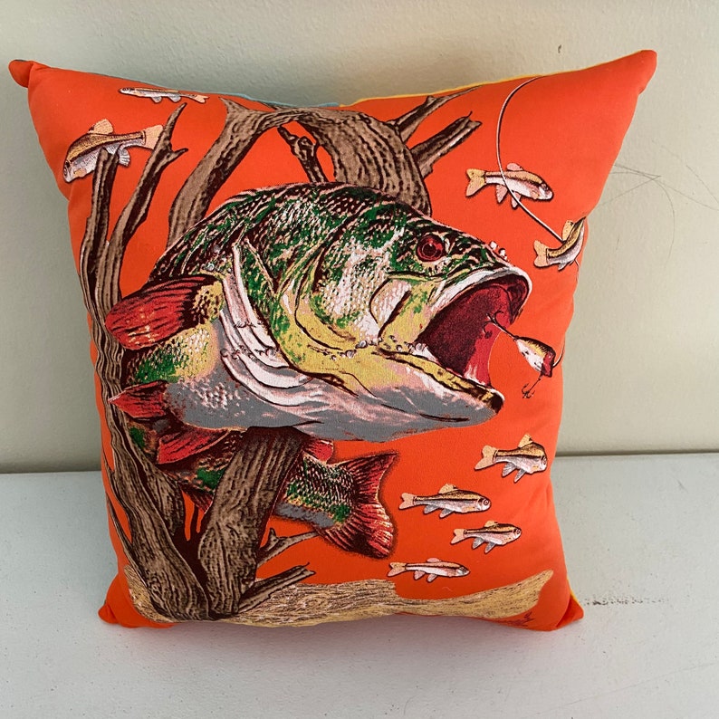K&ouml;nnte beinhalten: Ein orangefarbener Dekokissen mit einer gro&szlig;en Illustration eines Barsches mit offenem Maul. Der Fisch ist gr&uuml;n, braun und orange mit einem roten Maul. Kleinere orangefarbene Fische schwimmen um den Barsch herum.