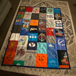 K&ouml;nnte beinhalten: Eine Patchwork-Decke aus verschiedenen T-Shirts. Die Decke ist blau, rot, wei&szlig;, gr&uuml;n, orange und grau. Die T-Shirts haben verschiedene Logos und Designs, darunter "BYU Cougars", "Padres", "Kindness", "Orlando USA", "DFSNS", "Little Monsters", "Century Football", "International" und "Mountain's Colorado".