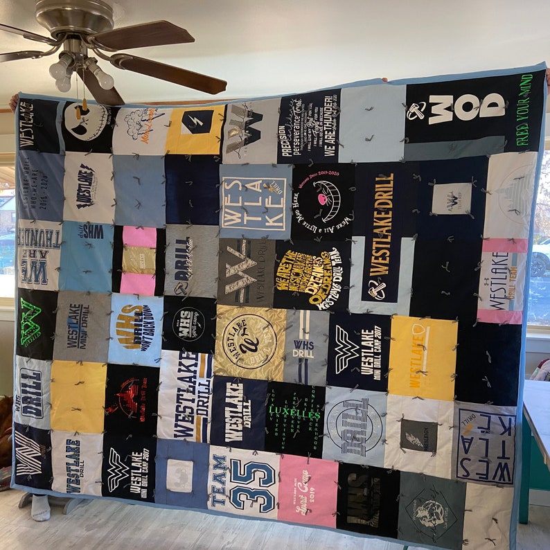 K&ouml;nnte beinhalten: Eine blaue und wei&szlig;e Decke aus T-Shirts mit den Worten "Westlake" und "Drill", die sich durchgehend wiederholen. Die Decke besteht aus Quadraten unterschiedlicher Gr&ouml;&szlig;e und Farbe. Die Decke h&auml;ngt an einer Wand.