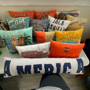 K&ouml;nnte beinhalten: Eine Sammlung bunter Kissen mit verschiedenen Designs und Texten, darunter "The Pound Clock", "Harley-Davidson", "I Got Hammered" und "America".