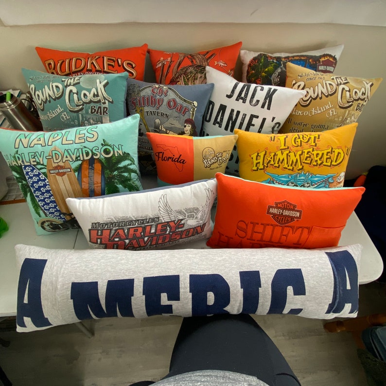K&ouml;nnte beinhalten: Eine Sammlung bunter Kissen mit verschiedenen Designs und Texten, darunter "The Pound Clock", "Harley-Davidson", "I Got Hammered" und "America".