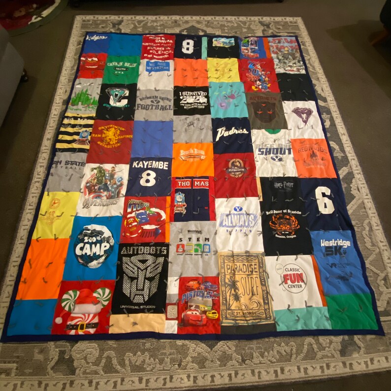 K&ouml;nnte beinhalten: Eine Patchwork-Decke aus verschiedenen T-Shirts. Die Decke ist blau, gr&uuml;n, rot, orange, gelb und wei&szlig;. Die T-Shirts haben verschiedene Logos und Texte, darunter "Kayembe 8", "Autobots", "Paradise Surf" und "Classic Fun Center".