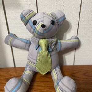 K&ouml;nnte beinhalten: Ein grauer und gr&uuml;ner karierter Teddyb&auml;r mit gr&uuml;ner Krawatte. Der B&auml;r sitzt auf einer Holzoberfl&auml;che.