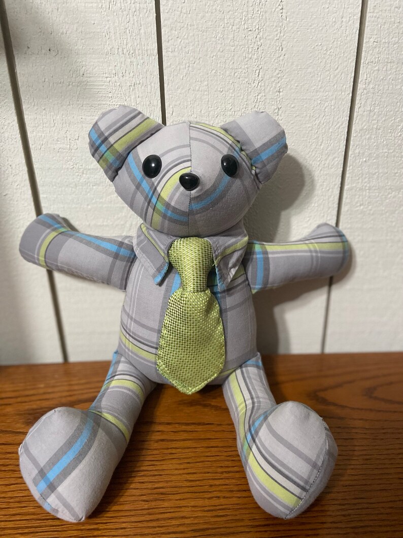 K&ouml;nnte beinhalten: Ein grauer und gr&uuml;ner karierter Teddyb&auml;r mit gr&uuml;ner Krawatte. Der B&auml;r sitzt auf einer Holzoberfl&auml;che.