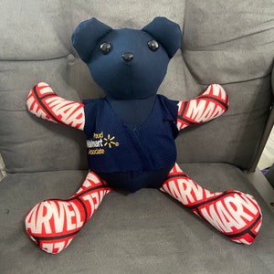 K&ouml;nnte beinhalten: Ein blauer Pl&uuml;schteddyb&auml;r, der eine blaue Weste mit dem Text "Proud Walmart Associate" und eine rote und wei&szlig;e Marvel-Hose tr&auml;gt.