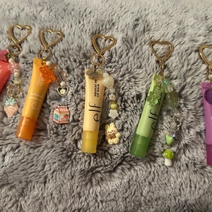 Puede incluir: Cinco tubos de bálsamo labial de colores con llaveros adjuntos. Cada tubo tiene la palabra "elf" y "Squeeze Me Lip Balm" impresa. Los llaveros están decorados con cuentas, dijes y pequeñas figuritas.