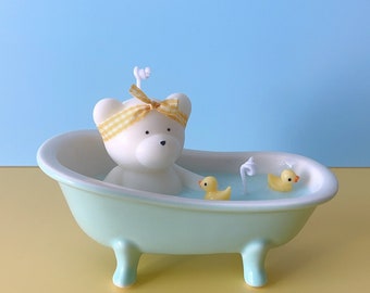 May include: A white ceramic bathtub with a white bear candle inside and two yellow rubber duck candles. The bathtub is light blue and has three legs.