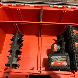 May include: A red toolbox with black dividers and multiple black battery packs. The battery packs are labeled "Milwaukee" and have a warning label. The toolbox has a clear plastic lid.